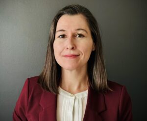 Portrait photograph of Dr. Mariel Herbert who is a pale woman with brown hair and some gray hair, brown eyes; she is wearing a white blouse and mulberry colored blazer and is smiling slightly.